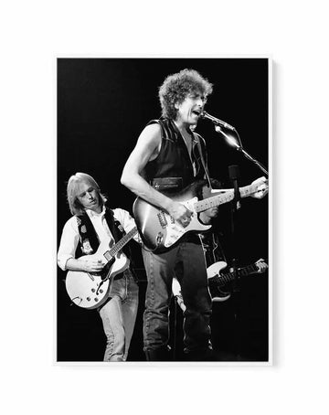 Framed black and white canvas print of musicians Bob Dylan and Tom Petty performing on stage. Bob Dylan, with curly hair and wearing a sleeveless vest, is singing into a microphone and playing an electric guitar in the foreground. Tom Petty, with long blonde hair and wearing a white shirt and vest, is playing an electric guitar slightly behind and to the left of Dylan. The background is dark, suggesting a concert stage, with another musician partially visible on the right.