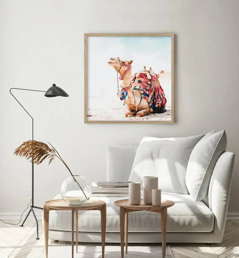Lifestyle shot of a minimalist living room featuring a framed art print of a bohemian camel above a white sofa. The print depicts a light brown camel adorned with a colourful, patterned blanket and blue and pink tassels, sitting on light sand under a pale blue sky. The print is displayed in a natural wood frame with a white border. The room includes a black floor lamp, two wooden side tables, a clear vase with dried pampas grass, and three cream-coloured ceramic vases.