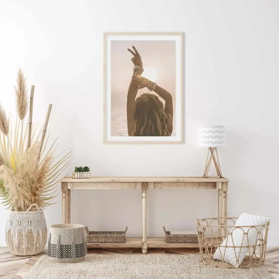 Lifestyle shot of a minimalist living room featuring a framed art print of a woman with her back to the camera, arms raised in a peace sign gesture, silhouetted against a bright sunset over the ocean. The print is displayed in a natural wood frame with a white border above a light wood console table. The room is decorated with dried pampas grass in a large woven basket, a smaller patterned basket, and a woven lamp.
