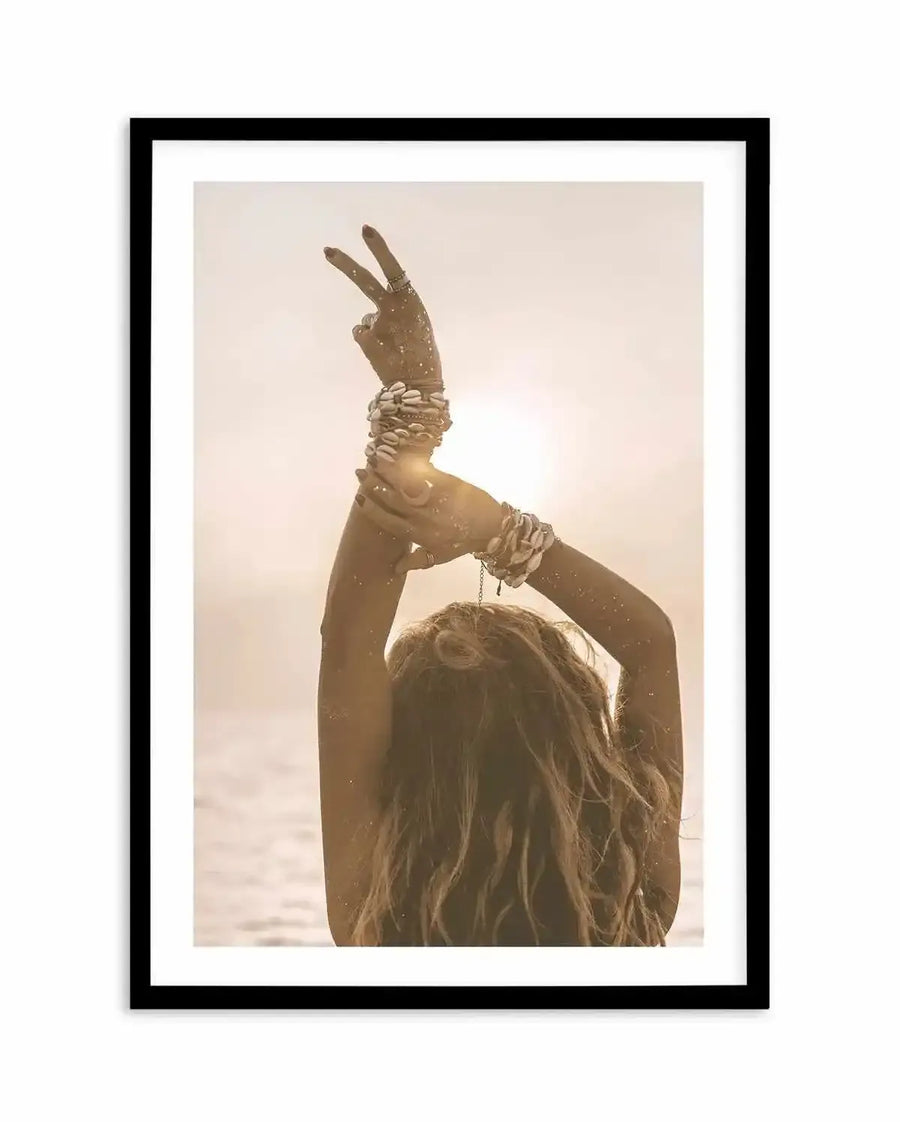 Framed photography art print featuring a bohemian-style woman with her back to the viewer, arms raised above her head, making a peace sign with one hand, against a bright, sun-drenched beach background. Her arms are adorned with multiple cowrie shell bracelets, and her long, wavy hair cascades down her back. The image has a warm, golden hour glow, printed on fine art paper and displayed in a black frame with white border.