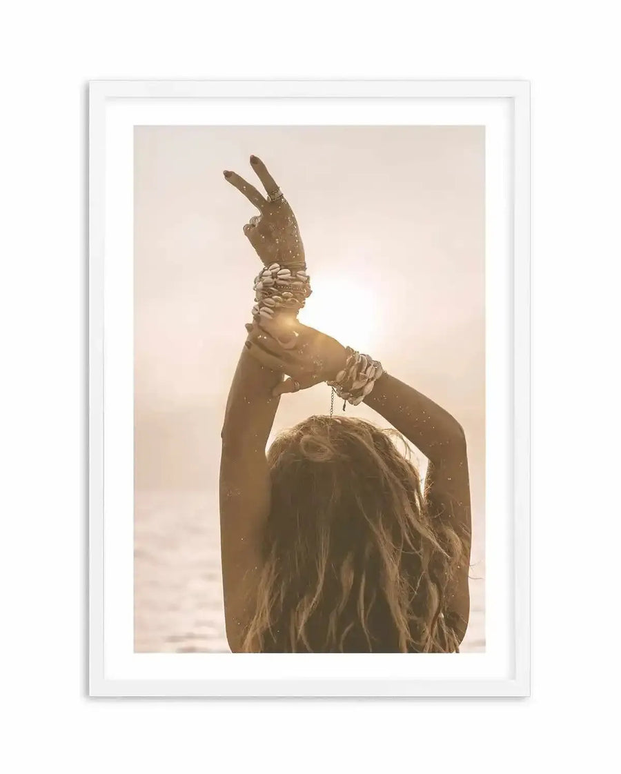 Framed sepia-toned photograph of a woman with her back to the camera, arms raised above her head, making a peace sign with her left hand. Her arms are adorned with multiple shell bracelets, and her long, wavy hair cascades down her back. The bright sun creates a warm glow behind her, with the ocean visible in the background, displayed in a white frame with a white border.
