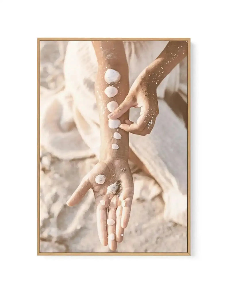 Framed canvas art print featuring two hands with golden glitter decoration holding white spherical objects arranged vertically along the arms and hands. Soft, warm, neutral tones with cream and beige sand-like background. Ethereal, dreamy aesthetic with flowing white fabric visible. Natural light creates soft shadows. Light wood-toned frame with white border surrounding the artwork.