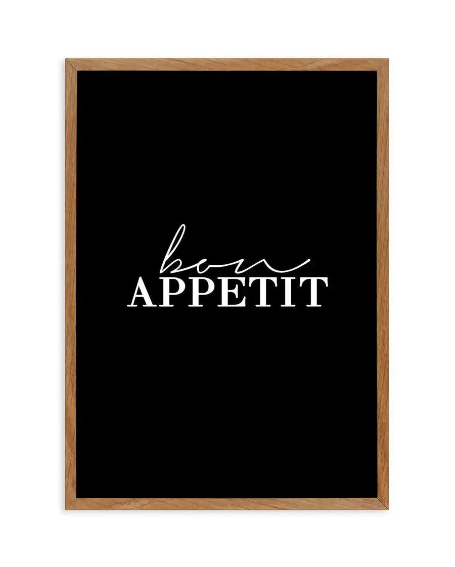 Framed art print with warm natural wood frame displaying black background with white cursive and sans-serif typography reading "bon APPETIT" in center. Cursive "bon" script flows above the word "APPETIT" in capital letters with a horizontal line underneath. Minimalist kitchen or dining room decor design with high contrast monochromatic color scheme.