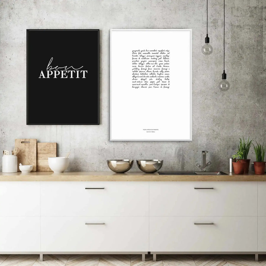 Lifestyle shot of a modern kitchen featuring two framed art prints on a concrete wall above a light wood countertop with white cabinets. The left print is a black art print with the words "bon APPETIT" in white script and sans-serif font, displayed in a black frame. The right print is a white art print with a list of cheese names in black script font, displayed in a white frame. Kitchen items include white ceramic bowls, cutting boards, stainless steel bowls, a sink with a chrome faucet, and potted plants.