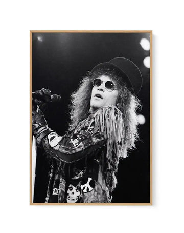 Framed black and white photograph of Bon Jovi frontman Jon Bon Jovi performing on stage, printed on canvas and displayed in a natural wood frame. The composition captures Bon Jovi mid-song, looking upwards with a microphone in his gloved hand. He wears a hat, round sunglasses, and a fringed jacket adorned with patches including a peace sign and skulls. The background is dark with blurred stage lights.