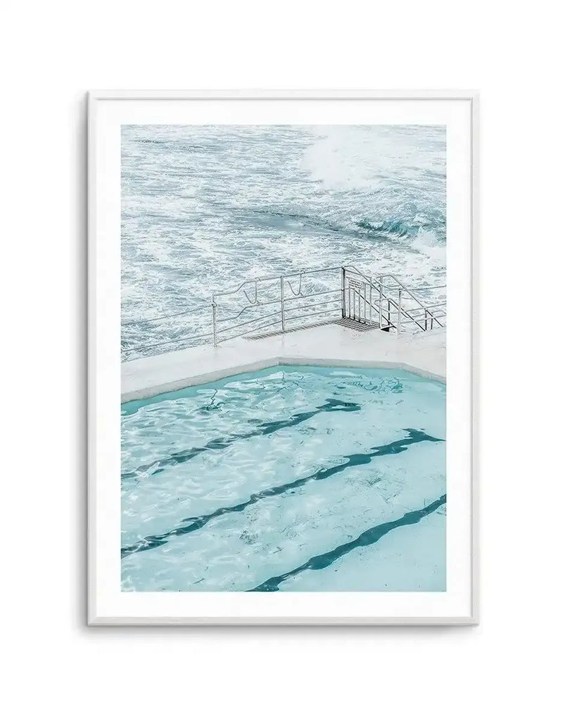 Framed art print featuring a photograph of an ocean-side swimming pool overlooking turquoise waves. The composition shows a light turquoise lap pool with dark lane dividers in the foreground, a metal railing fence running across white concrete in the middle ground, and turbulent grey-green ocean waves with white foam in the background. The photograph has a soft, muted color palette dominated by aqua, seafoam, and grey-blue tones. The image is presented in a silver-toned frame with a white border.