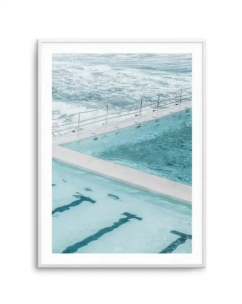 Framed art print showing an aerial view of an ocean swimming pool divided into three sections. The foreground displays a pale turquoise pool with dark lane dividers visible underwater. The middle section shows a deeper teal pool separated by white concrete edging, with a metal safety railing. The background features turbulent ocean waves in shades of blue and white beyond the railing. The photograph has cool aquatic tones throughout. The print is displayed in a light gray wooden frame with a white border.