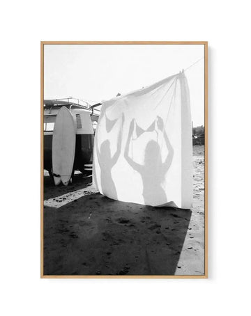 Black and white framed canvas art print titled "Born Free" by Mario Stefanelli, displayed in a natural wood frame. The photograph shows a white fabric or sail billowing in the wind on a sandy beach, with silhouettes of two figures visible through the translucent material. A vintage vehicle and surfboard are positioned to the left. The composition captures movement and freedom against a bright sky, printed on canvas with a white border, mounted in a warm tan wooden frame.