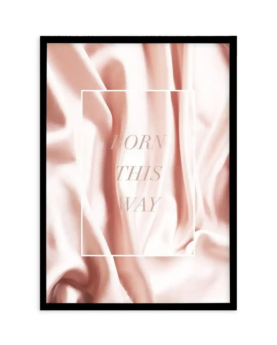 Art print featuring a close-up of flowing, draped fabric in a soft, muted blush pink with subtle highlights and shadows, creating a silky texture. The words "BORN THIS WAY" are printed vertically in a light, elegant serif font within a thin white rectangular border in the center of the print, displayed in a black frame with a white border.