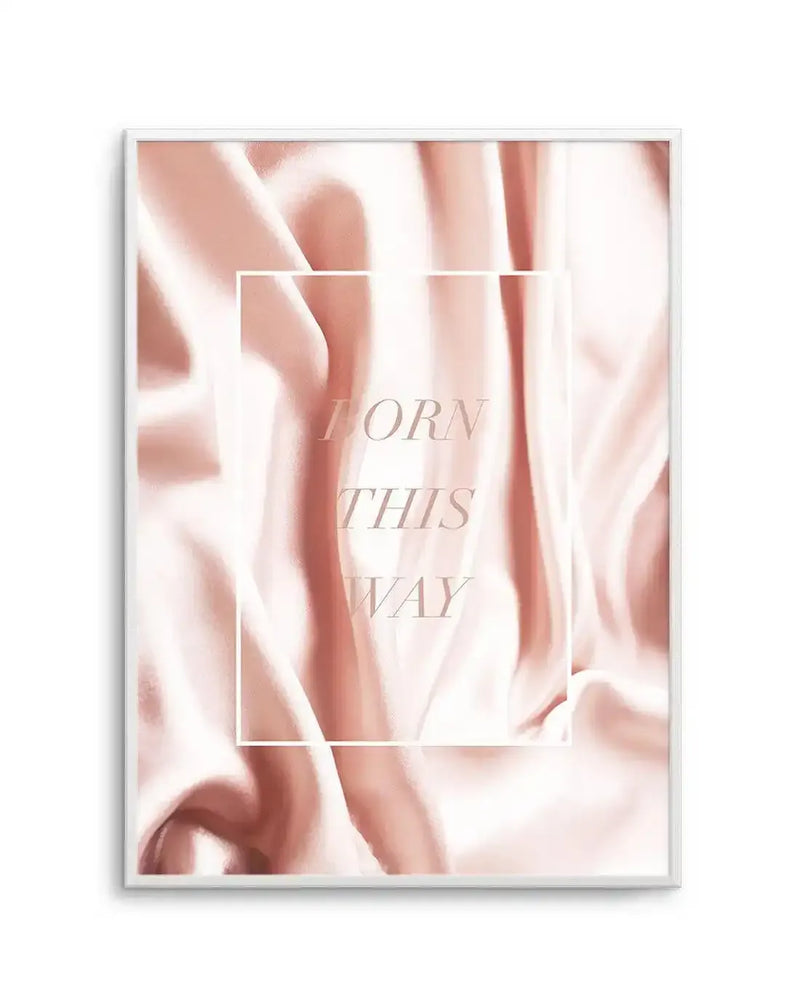 Art print featuring a close-up of flowing, draped fabric in shades of blush pink and soft cream, creating a silky, luxurious texture. The words "BORN THIS WAY" are subtly overlaid in a light pink serif font, enclosed within a delicate white rectangular frame. The print is displayed in a thin white frame.