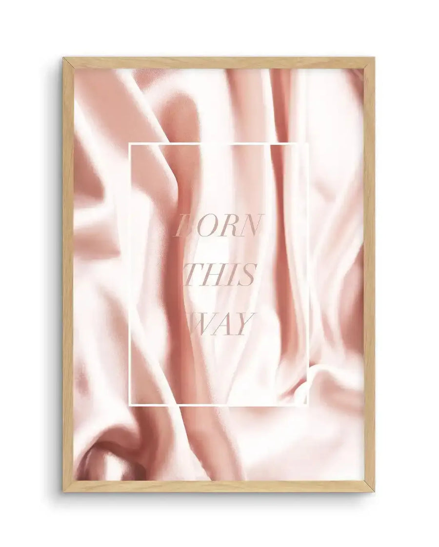 Art print featuring a pale pink and cream satin fabric with soft folds and drapes, with the words "BORN THIS WAY" in light pink capital letters centered in a white rectangular outline, displayed in a natural oak frame with white border.