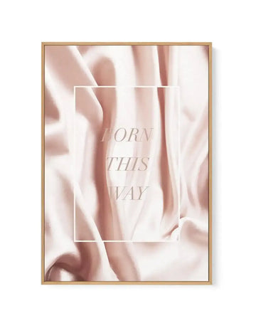 Framed canvas art print with a natural wood frame displaying soft, blurred abstract imagery in blush pink and cream tones. The composition features flowing, organic shapes suggesting draped fabric or petals. Centered within a white border is the text "BORN THIS WAY" in elegant, light gray serif lettering. The overall aesthetic is serene and minimalist with a soft-focus photographic quality.