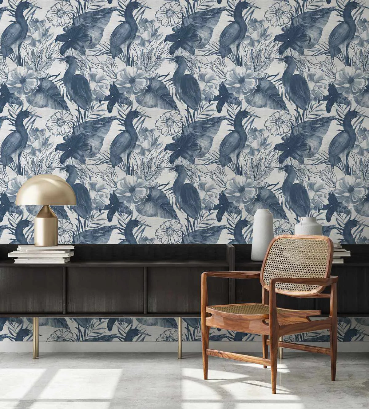 Botanica in Navy Wallpaper displayed on a room wall featuring a repeating pattern of blue-grey herons and birds among watercolor-style flowers including dahlias and daisies on a cream background. The wallpaper is shown in a styled interior with a black modern sideboard, brass mushroom table lamp, white ceramic vessels, stacked books, and a wooden mid-century chair with woven rattan backing positioned in front of the patterned wall.