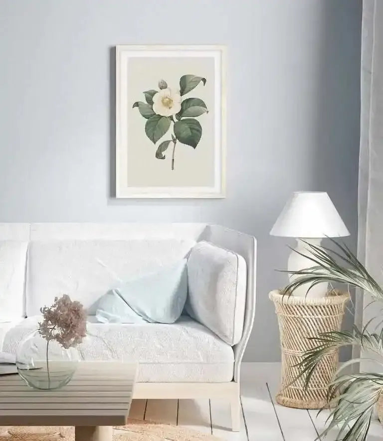 Large framed art print displayed above a curved white sofa in a minimalist living room. The print features a white camellia flower with a yellow center and dark green leaves on a light cream background, with a natural wood frame and white border. The room includes a light blue throw pillow, a wicker side table with a white lamp, and a potted plant with long green leaves.