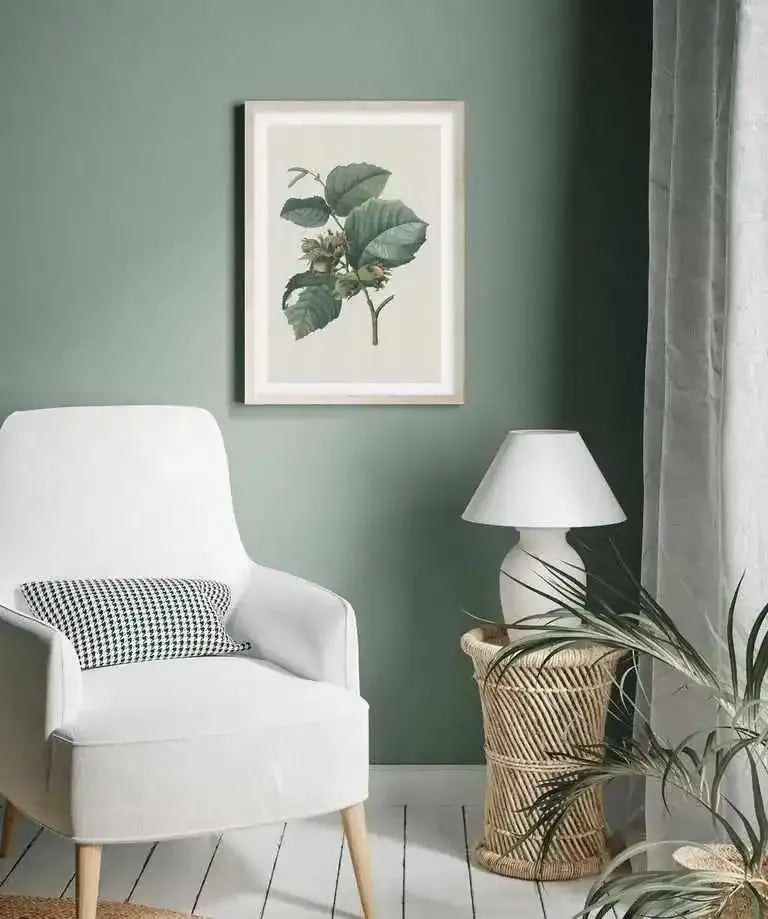 Lifestyle shot of a minimalist living room with a framed art print displayed on a sage green wall above a white armchair. The art print features a botanical illustration of a hazelnut branch with large green leaves and clusters of unripe hazelnuts, set against a cream background with a natural wood frame and white border. A white table lamp sits on a woven rattan side table next to the armchair.