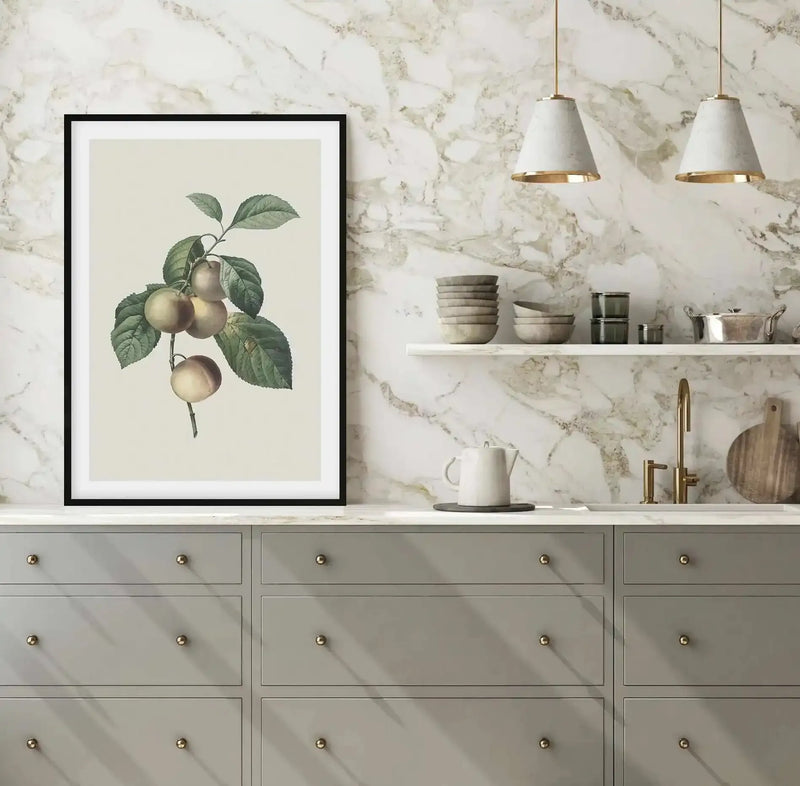 Lifestyle shot of a modern kitchen featuring a framed art print of a botanical illustration. The print depicts a branch with several round, light green fruits and dark green leaves against a pale cream background, displayed in a black frame with a white border. The kitchen has white marble backsplash, light grey cabinets with gold knobs, a white countertop, a gold faucet, and two white pendant lights with gold accents.
