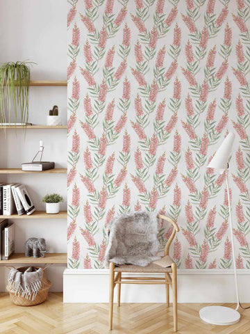 Bottlebrush Flowers in Red Wallpaper displayed on a bedroom wall featuring a repeating pattern of dusty rose and coral bottlebrush flowers with sage green foliage on a cream background. The botanical illustration style wallpaper is shown installed behind a natural wood chair with gray sheepskin throw, alongside wooden floating shelves holding books, plants, and decorative objects on a herringbone wood floor.