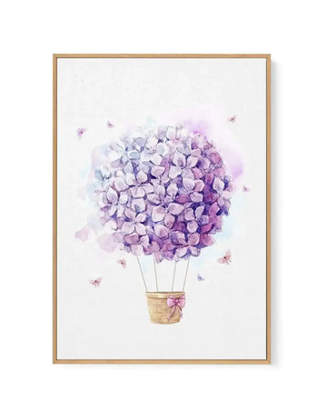 Watercolour illustration of a hot air balloon made from a large spherical cluster of purple and lavender hydrangea flowers, suspended above a woven wicker basket tied with a pink ribbon. Tiny purple and mauve butterflies flutter around the balloon against a soft white background with subtle watercolour washes. The artwork is printed on canvas and set in a natural wood frame with a white border.