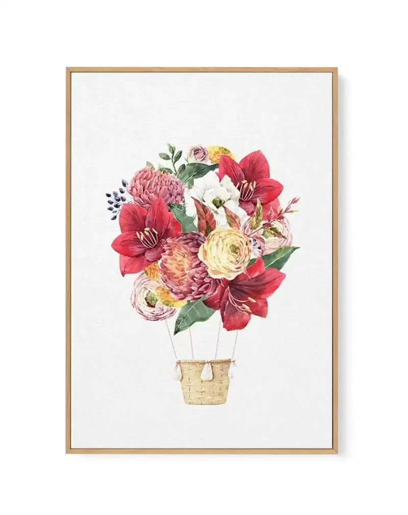 Watercolor illustration of a hot air balloon made entirely of vibrant flowers in shades of coral red, dusty rose, pale yellow, mauve, and cream, with green foliage and stems, suspended from a woven tan basket. The composition is centered on a white background, framed in natural wood with a white border.