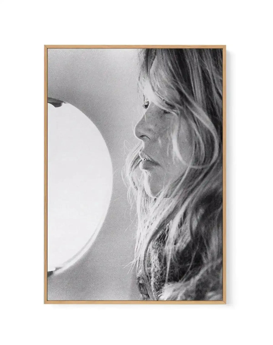 Black and white photographic portrait of a man in profile facing left, framed in natural wood frame. The subject has long, wavy hair and a full beard, gazing downward with a contemplative expression. A large white circular form, possibly a moon or light source, is visible on the left side of the composition. The photograph has a grainy, vintage quality typical of mid-20th century film photography. The natural wood frame has a warm tan finish and is displayed against a white background.