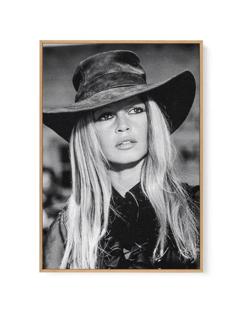Black and white framed canvas art print featuring a portrait of a woman wearing a wide-brimmed cowboy hat and dark shirt. The photograph is a close-up headshot with long straight blonde hair, defined eye makeup, and a neutral expression. The image has high contrast grayscale tones typical of vintage portrait photography. The canvas is mounted in a natural wood frame with a white border around the photograph.