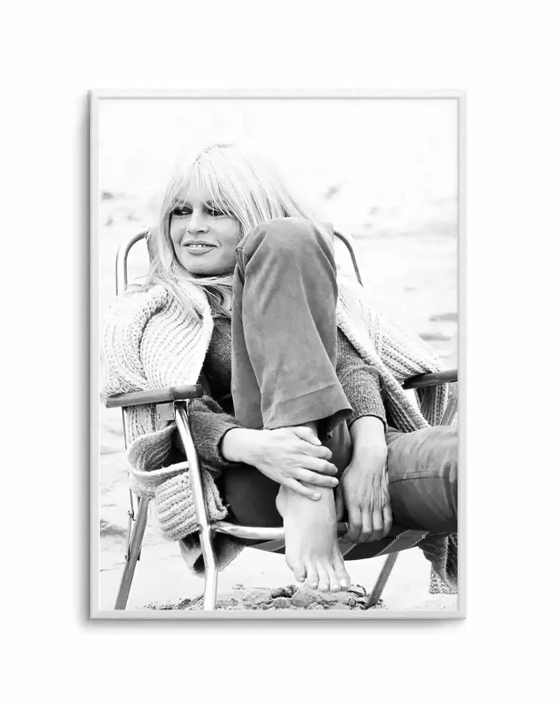 Framed black and white photograph of Brigitte Bardot sitting in a beach chair on sand, with a natural wood frame and white border. She is looking to the left with a slight smile, wearing a light-coloured knitted cardigan and dark trousers, with her left leg bent and her bare foot resting on her right leg.