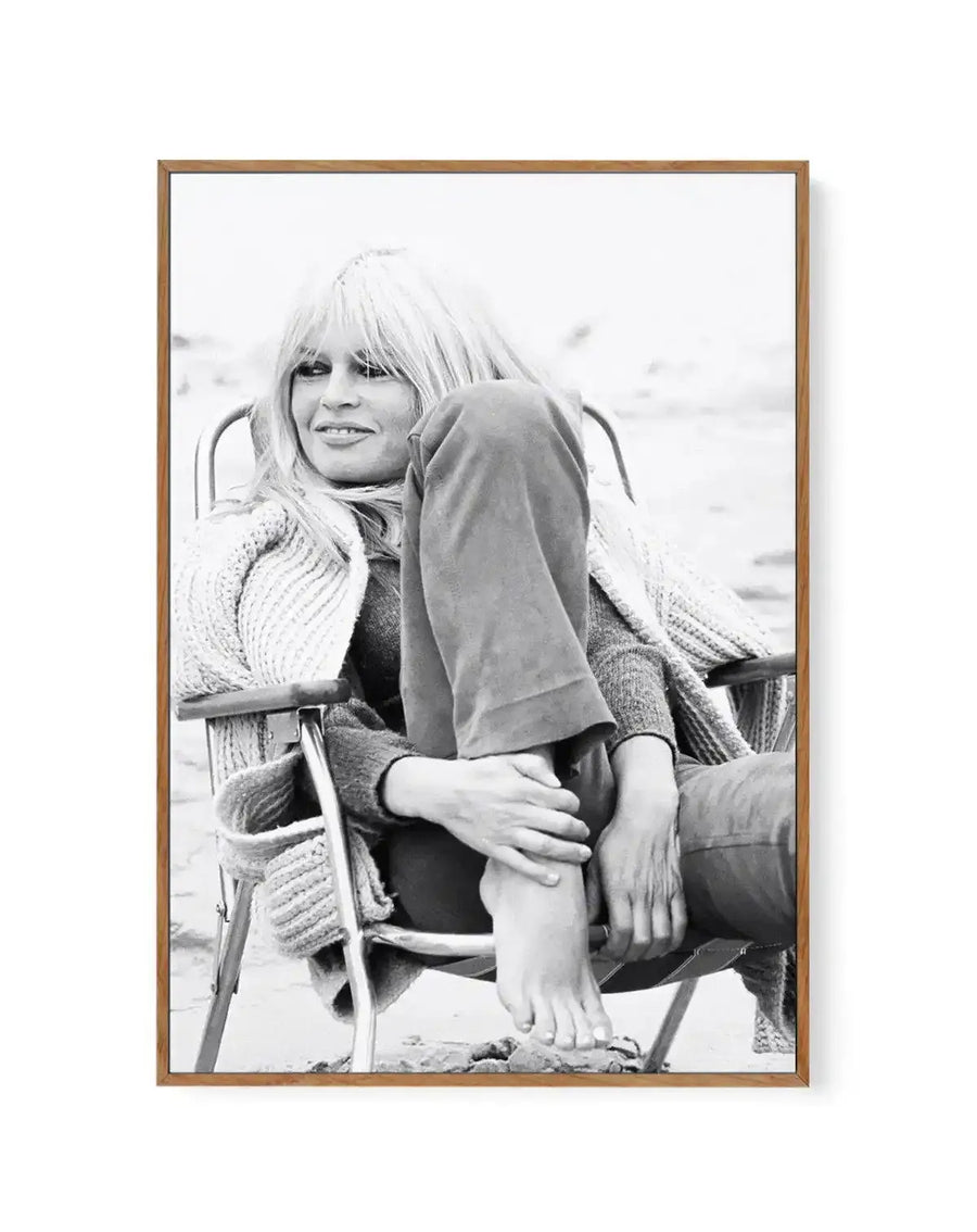 Black and white framed canvas art print featuring a woman with blonde bangs seated in a chair, smiling at the camera. She wears a textured knit cardigan over a dark top and poses with one leg extended. The photograph has a casual, candid quality with a blurred background. The canvas is mounted in a natural wood frame with a white border.