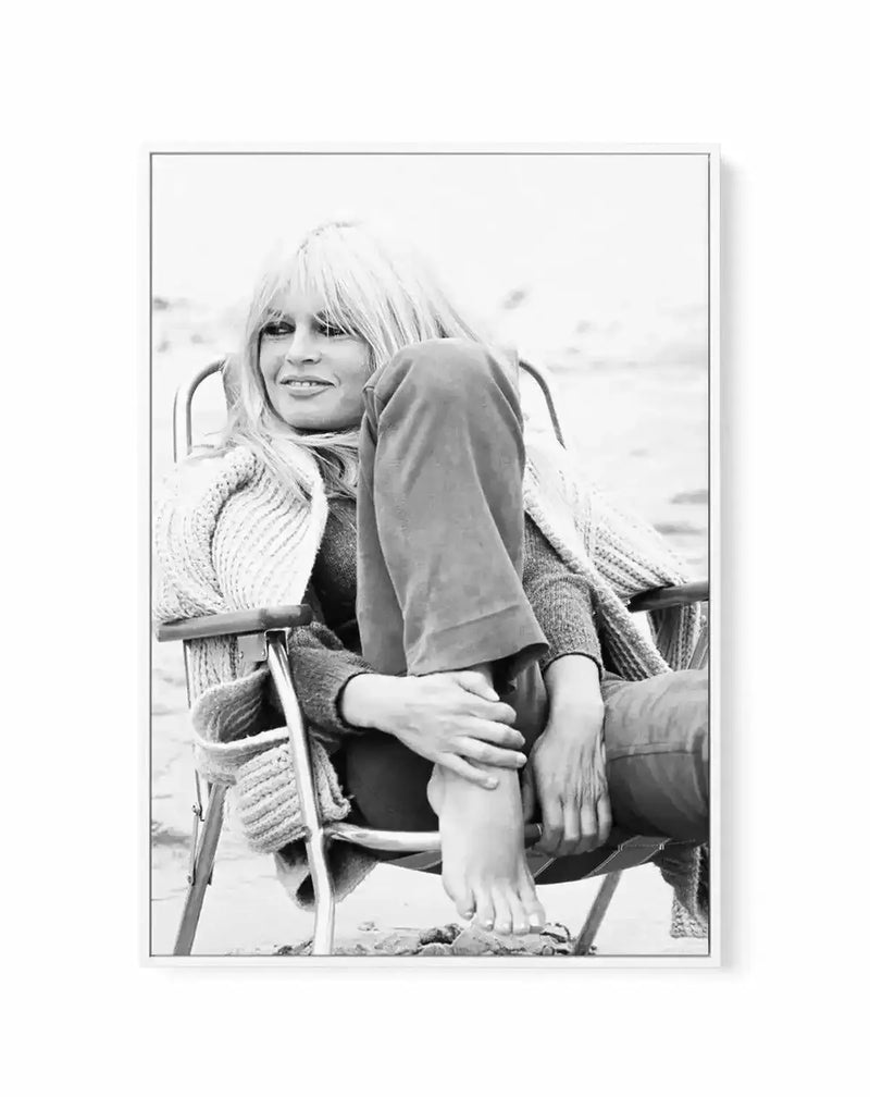 Black and white framed canvas art print featuring a classic photograph of a woman with blonde bangs seated in a chair, smiling at the camera. She wears a knitted cardigan over dark clothing and poses with one leg extended. The photograph has a soft, vintage quality with a blurred background suggesting an outdoor setting. The canvas is mounted in a gray frame with a white border.