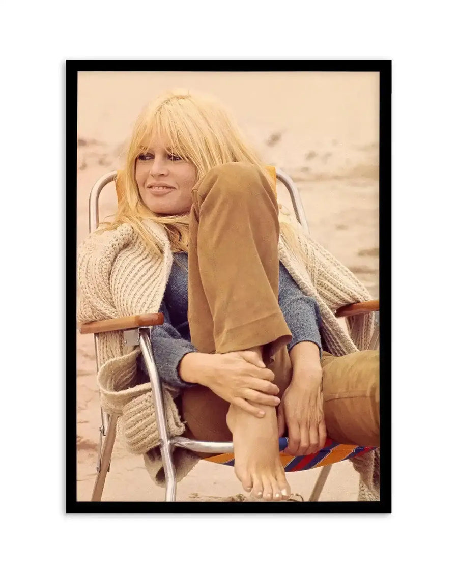 Framed full-colour photography art print featuring Brigitte Bardot sitting on a beach chair. Bardot, with long blonde hair and bangs, is looking to the left with a slight smile, wearing a blue top, brown trousers, and a cream knitted cardigan. Her right foot, with white painted toenails, is resting on her left knee. The background is a sandy beach with a light beige sky. The print is displayed in a black frame with a white border.