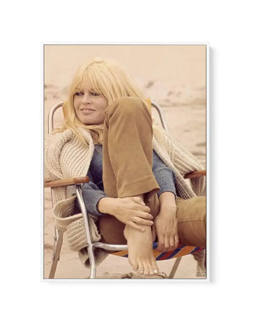 Framed canvas art print featuring a color photograph of a woman with blonde bangs seated in a beach chair on sand. She wears a blue-grey knit sweater and cream cable-knit cardigan, with a tan fabric draped across her legs. The photograph has warm, muted tones dominated by beiges, tans, and soft blues. The canvas is mounted in a natural wood frame with a white border.