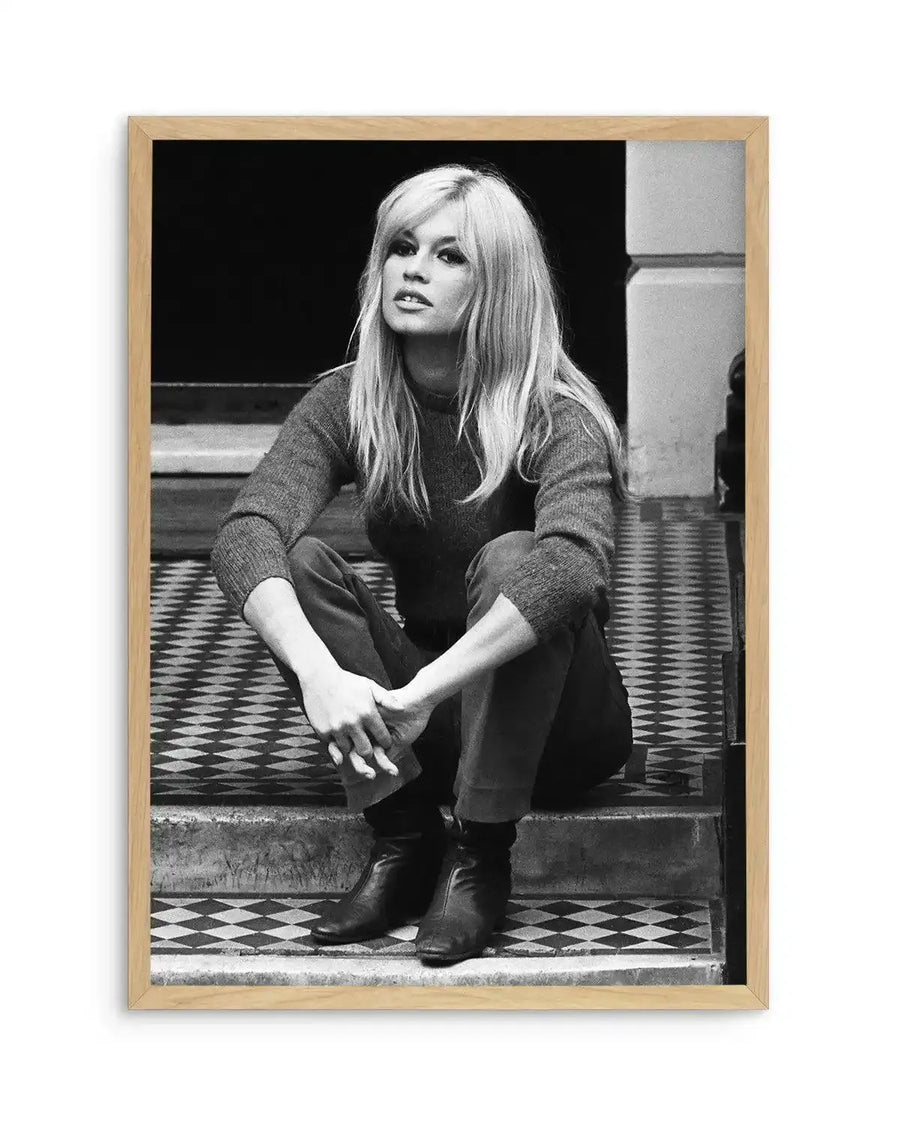 Framed black and white photograph of Brigitte Bardot sitting on a tiled stoop, with natural wood frame and white border. The composition features Bardot in a dark sweater and pants, with long blonde hair and bangs, looking directly at the camera with a slightly open mouth. She is sitting with her knees bent and hands clasped, wearing dark boots. The background shows a dark doorway and a patterned tile floor.