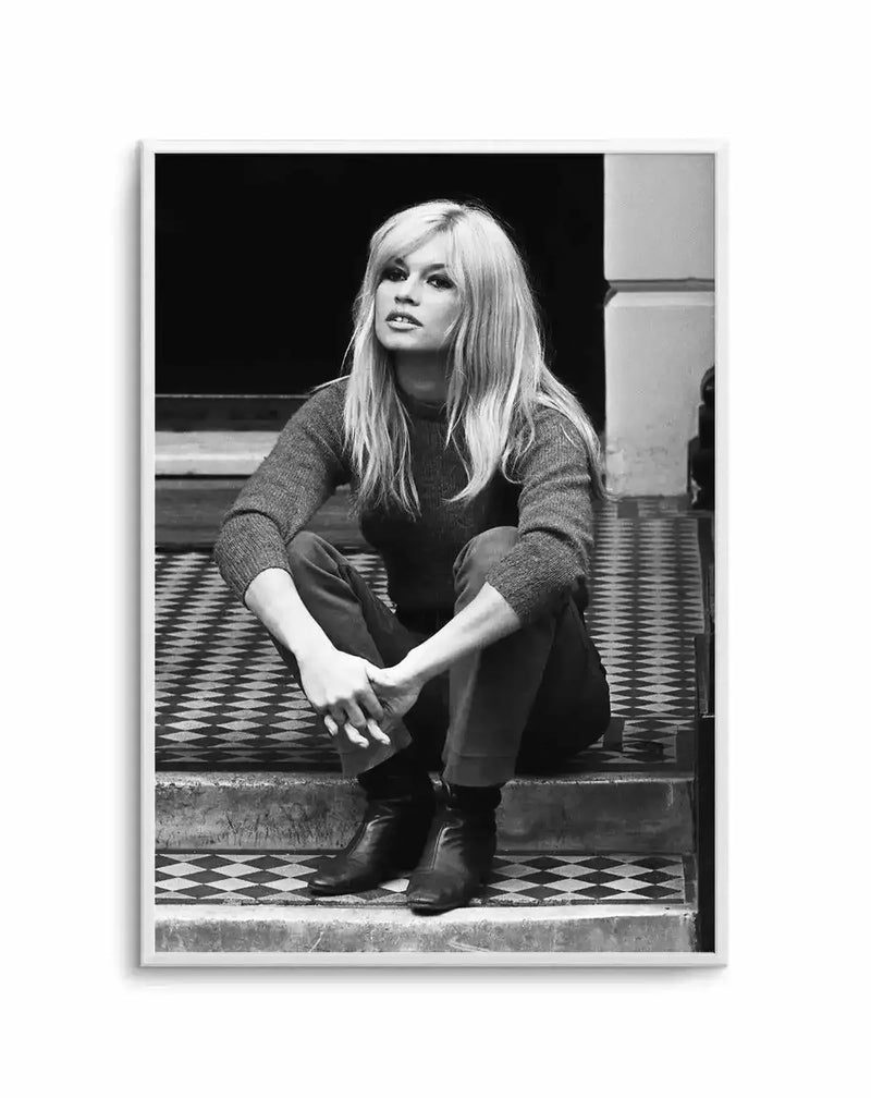 Framed black and white photograph of Brigitte Bardot sitting on tiled steps, with a natural wood frame and white border. The composition features Bardot with long blonde hair, wearing a dark sweater and trousers, looking slightly to the left with a thoughtful expression. Her hands are clasped in front of her, and she wears dark boots. The background is dark, with architectural elements visible on the right.