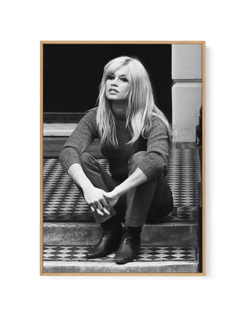 Black and white framed canvas art print featuring a woman with long blonde hair in 1960s style, seated on tiled steps wearing a dark ribbed sweater and dark pants. She gazes upward thoughtfully with her hands clasped near her knees. The photograph has high contrast black and white tones, mounted in a natural wood frame with white border, set against a minimalist interior backdrop.
