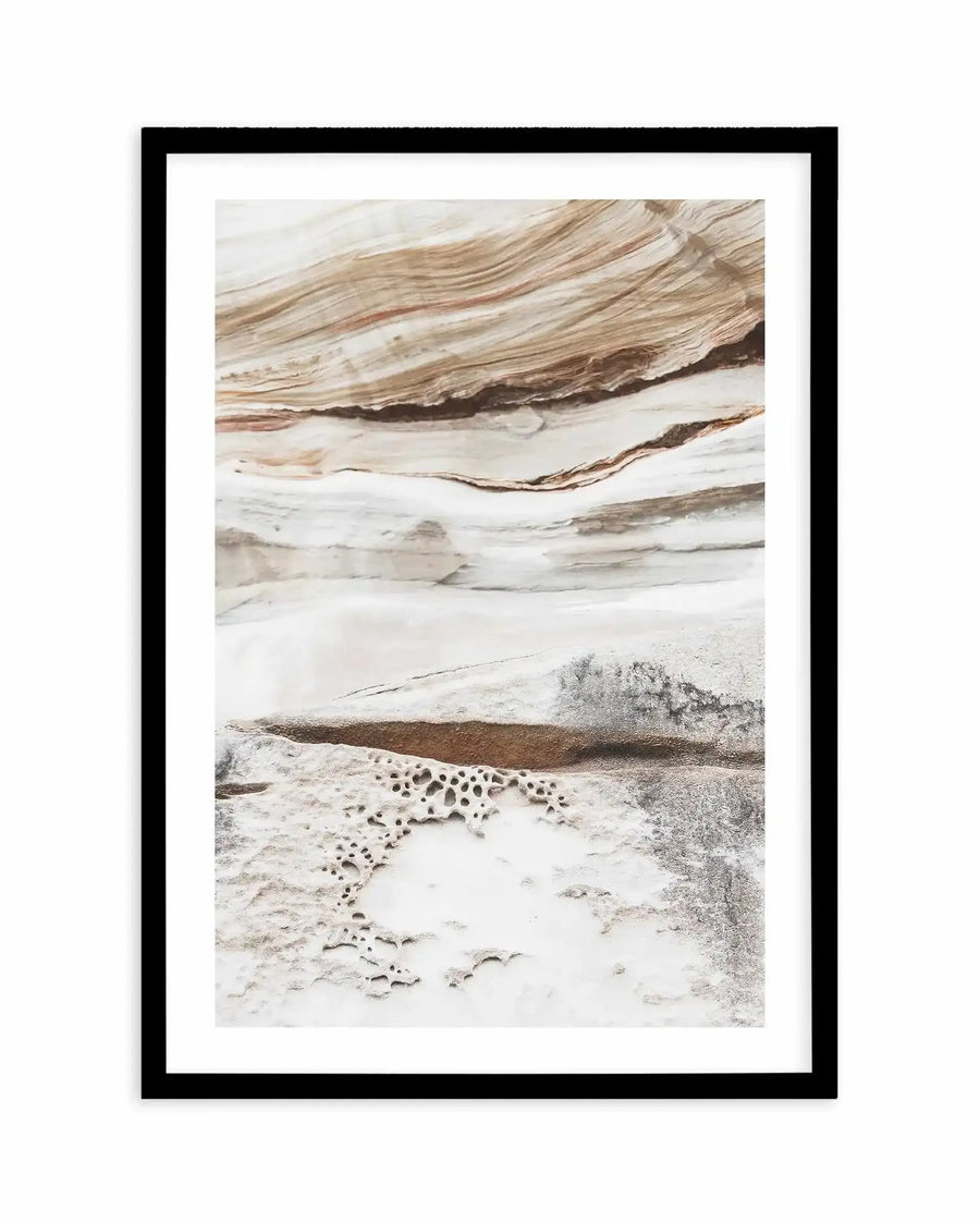 Framed art print titled Bronte Rocks III displayed against a white wall. The print features a minimalist photograph of layered rock formations in muted earth tones—warm tan, beige, cream, and soft brown. Horizontal striations create flowing patterns across the upper portion, while the lower section shows pale mineral deposits with dark circular holes or thermal features. The composition emphasizes natural textures and subtle color gradations. The print is mounted in a black frame with a white border.