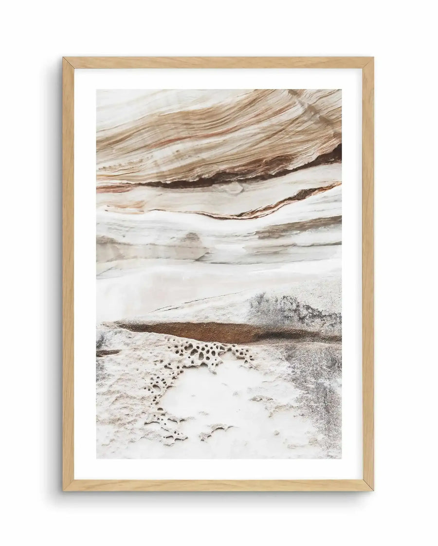 Framed art print featuring a photograph of layered rock formations in warm earth tones. The composition shows horizontal bands of cream, tan, and brown striations with natural texture and depth. Lower portion depicts mineral pools with circular holes and white crusty deposits against grey sand. Natural light wood frame with white border. Soft, neutral colour palette evokes geothermal or desert landscape geology.