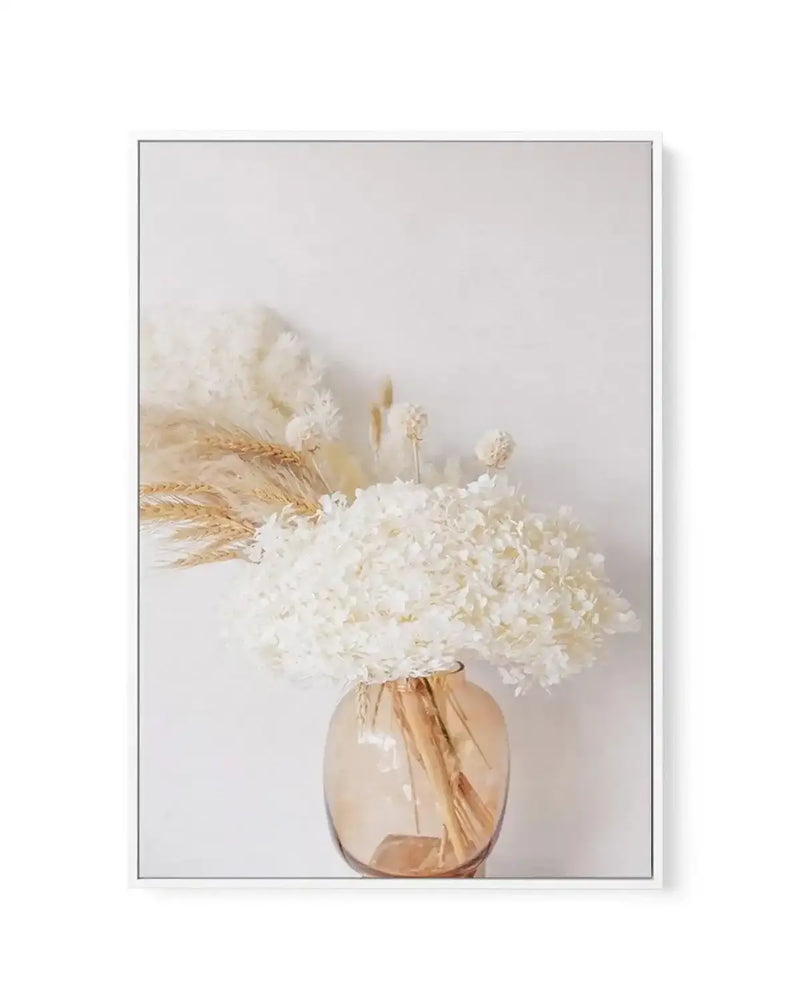 Framed canvas art print featuring a soft, photorealistic still life composition. A clear glass vase with warm bronze and amber tones holds a generous arrangement of white hydrangeas with dried wheat stems in golden tan. The vase sits centered against a soft, neutral beige-grey background. The canvas has a thin grey frame with a white border. The overall aesthetic is minimalist and calming with muted, natural tones throughout.