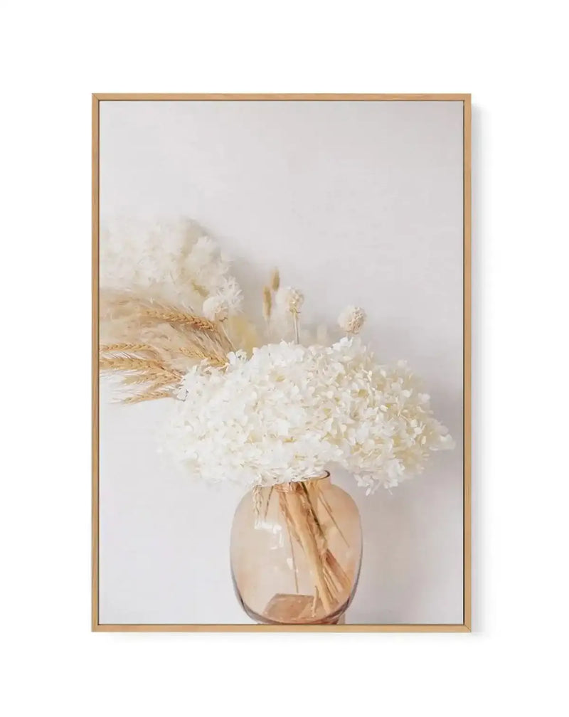 Framed canvas art print featuring a clear glass vase with warm peachy-bronze tones containing a lush arrangement of white hydrangeas and dried wheat stems in golden tan. The composition is centered against a soft, neutral white background with subtle warm undertones. The vase sits at the bottom of the vertical rectangular frame, which has a natural wood-tone border in light tan oak finish. The overall aesthetic is minimalist and botanical, with soft, muted colors and a calm, serene mood.