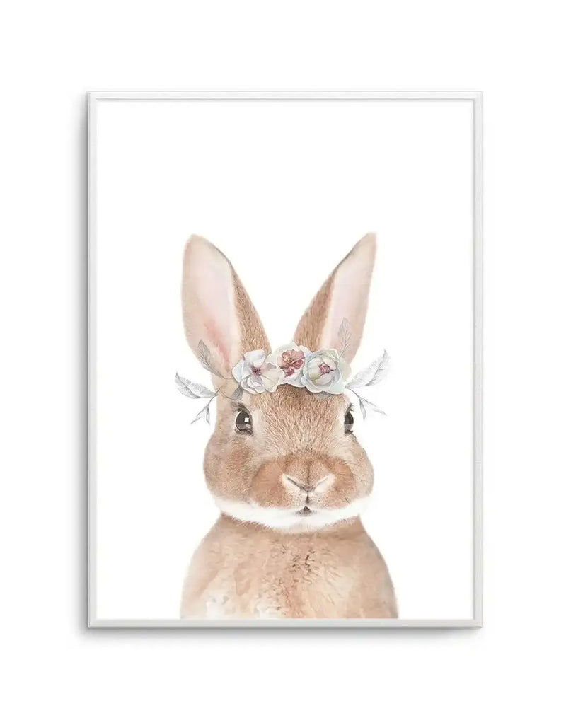 Watercolour art print featuring a fluffy brown bunny with a delicate floral crown on its head, displayed in a white frame with a white border. The bunny has soft brown fur, long ears with pink interiors, and a small pink nose, looking directly at the viewer with a gentle expression. The flower crown consists of small, muted pink, cream, and blue flowers with delicate grey-green leaves.