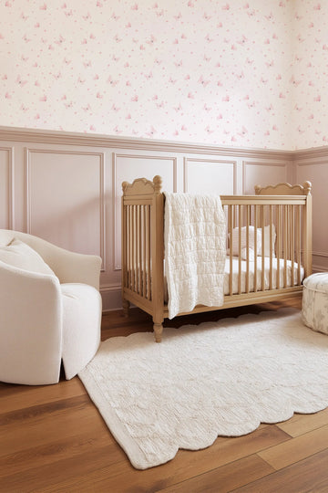 Lifestyle shot of a baby's nursery with light pink butterfly wallpaper and pale pink wainscoting. The wallpaper features a repeating pattern of delicate, translucent pink butterflies and small pink dots on a cream background. A light wood crib with a white quilted blanket draped over the side is in the center, with a cream boucle armchair to the left and a patterned ottoman to the right. A cream scalloped rug lies on the light wood floor.