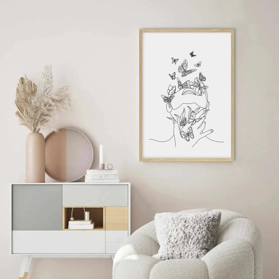 Large framed art print displayed above a curved white sofa in a minimalist living room. The print features a continuous line drawing of a woman's face with numerous butterflies emerging from her head and hands, rendered in black on a white background, with a natural wood frame and white border. The room includes a textured cream throw, dried pampas grass in a tall beige vase, and a two-toned round decorative tray.