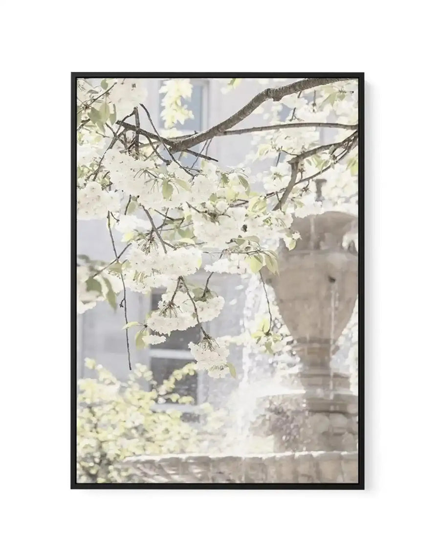 Framed canvas art print featuring delicate white flowering branches in soft focus in the foreground, with a blurred London fountain and classical architecture visible in the background. The composition uses a soft, muted palette of cream, pale yellow, sage green, and warm grey tones, creating a dreamy, impressionistic aesthetic. The photograph-style artwork is mounted in a black frame with white border, presented vertically against a white wall.