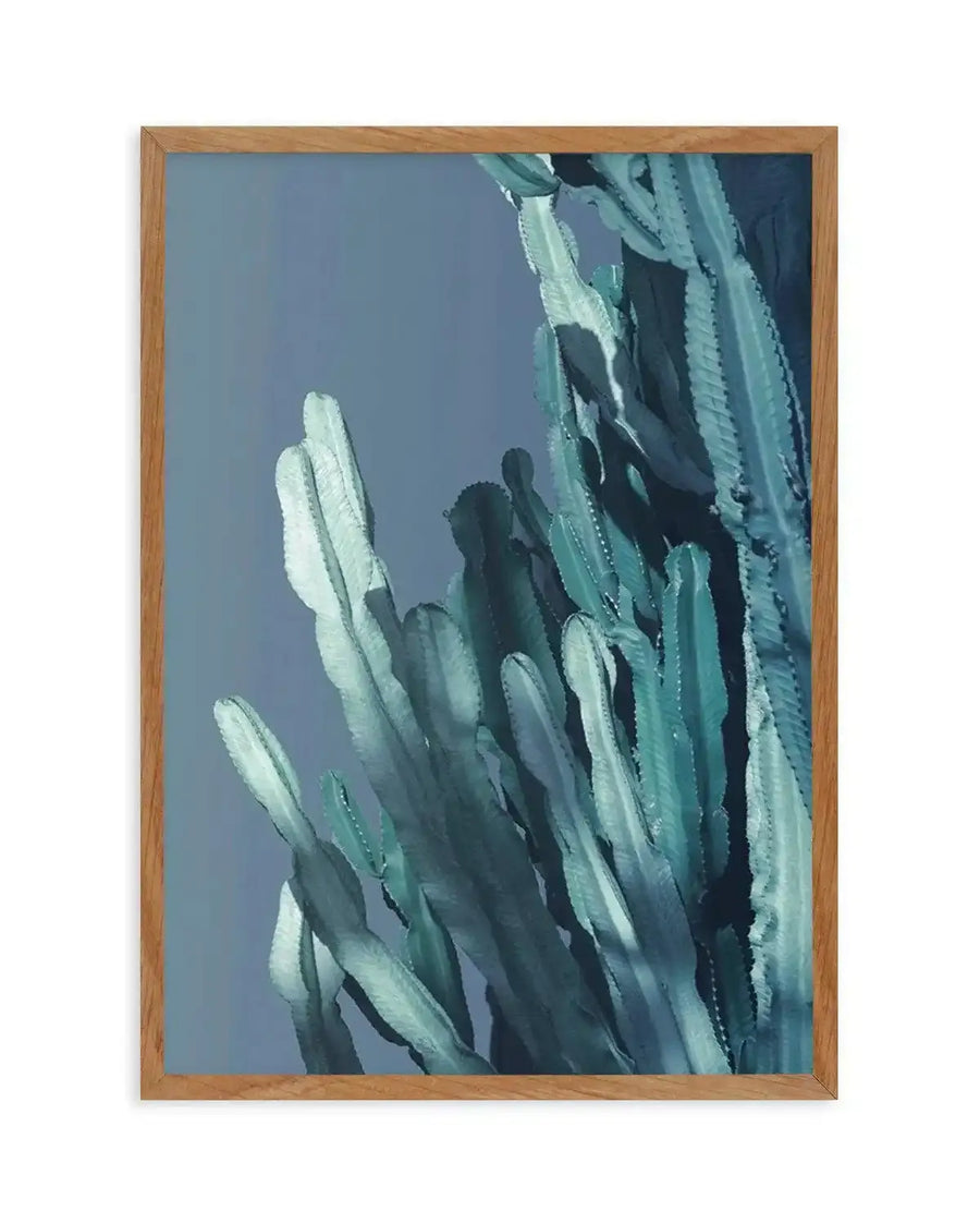 Framed art print featuring an abstract painting of tall cactus forms rendered in soft greens, teals, and whites against a muted blue-gray background. The composition uses loose, gestural brushstrokes creating a watercolor-like effect with areas of light and shadow that suggest dimension and movement. The wooden frame is natural tan oak. Vertical orientation emphasizing the upright growth of the plant forms.