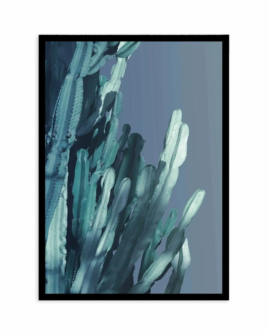 Framed art print featuring a close-up illustration of tall columnar cacti against a soft periwinkle-blue sky. The cacti have elongated, finger-like segments in varying shades of sage green, seafoam, and cream, with visible texture lines suggesting spines. The composition shows the plants from a low angle, emphasizing their vertical growth. The painting style appears watercolor or gouache with soft, blended edges and muted tones. Black frame with white border.