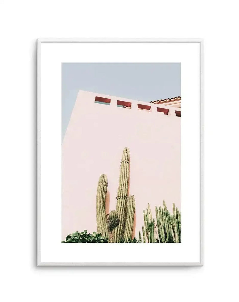 Framed art print featuring a tall saguaro cactus with smaller cacti beside it against a soft blush pink wall of a Southwestern-style building with red window openings and terracotta roof tiles visible at top. Clear blue sky above. Photograph printed on white paper with white border, displayed in a light gray frame.