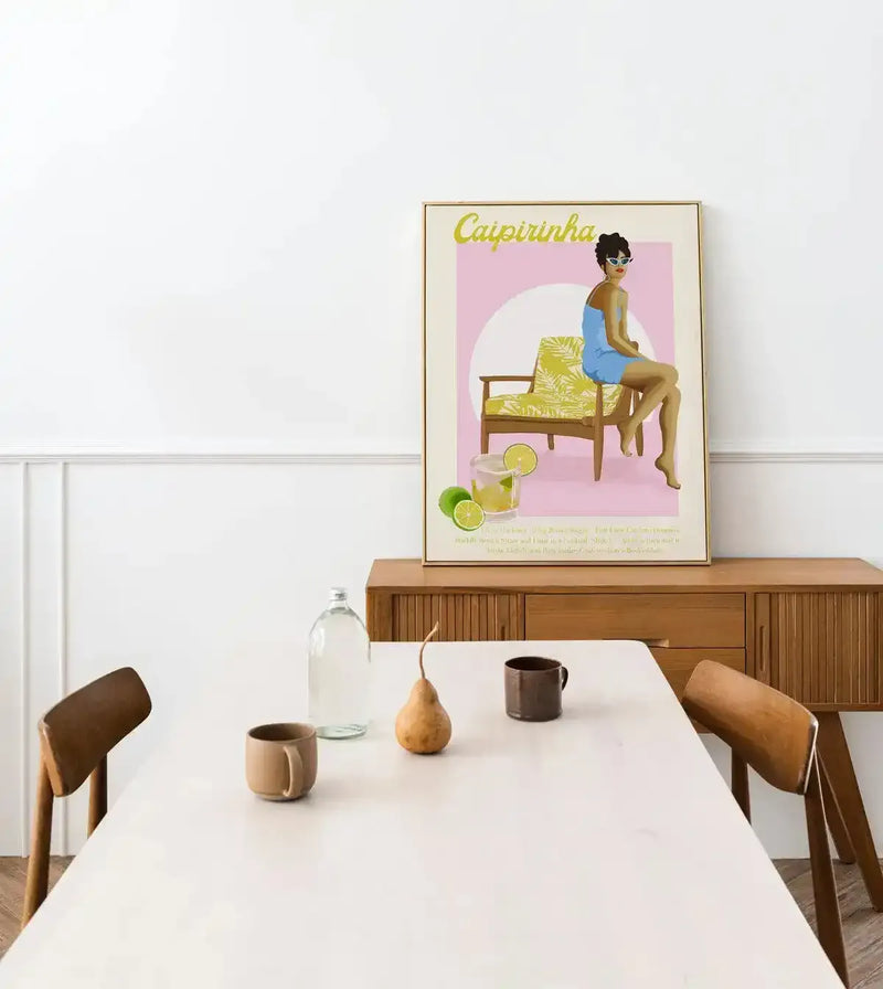Lifestyle shot of a Caipirinha art print displayed in a minimalist dining room. The print features a woman in a blue swimsuit and sunglasses sitting on a patterned chair, with a Caipirinha cocktail and limes in the foreground, all against a pink and cream background. The print has a natural wood frame and white border and is placed on a wooden buffet. A white dining table with two wooden chairs, a glass bottle, two mugs, and a decorative pear are in the foreground.