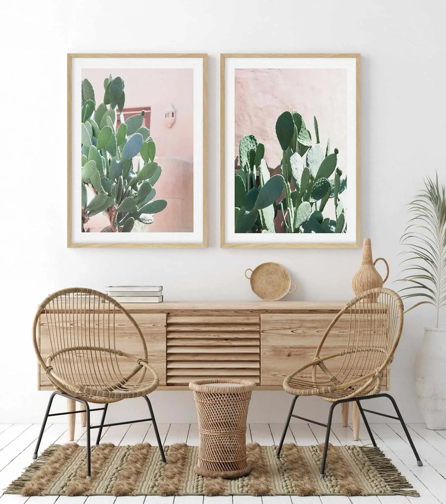 Framed art print of a prickly pear cactus with broad green paddle-shaped leaves against a soft blush pink background. Natural wood frame with white border. Displayed on white wall above wooden console table between two woven rattan chairs, styled with decorative baskets and potted plants in a minimalist bohemian interior setting.