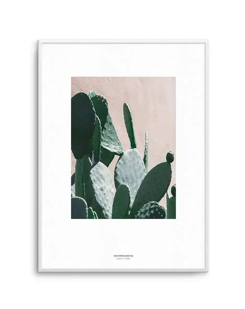 Framed art print featuring a close-up photograph of California prickly pear cactus against a soft beige wall. The composition shows multiple cactus paddles in varying shades of sage green and muted olive, with some displaying pale grey-green coloring and visible spines. The photograph has a calm, muted aesthetic with soft natural lighting. The print is mounted in a white frame with a white border, and includes small text at the bottom reading "CALIFORNIA CACTUS" and "DULCE ET CRUEL".