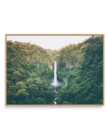 Framed canvas art print of Cannabullen Waterfall featuring a tall, slender waterfall cascading down a lush green valley surrounded by dense forest vegetation. The composition shows the waterfall in the center with steep, forested cliff walls on either side covered in varying shades of green. A white stream flows down the rocky base below the falls. The sky above is overcast and pale. The canvas is mounted in a natural wood frame with a light tan finish.