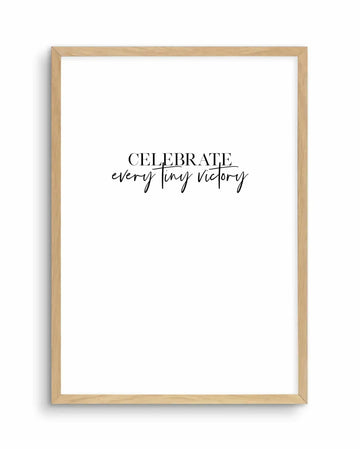 Art print in light natural wood frame displaying motivational text on white background. The design features "CELEBRATE" in serif capital letters, with "every tiny victory" written below in flowing cursive script. Black text on white creates a minimalist, modern aesthetic. The wooden frame has a warm honey tone with visible grain texture. The composition is centered and simple, suitable for home or office décor.