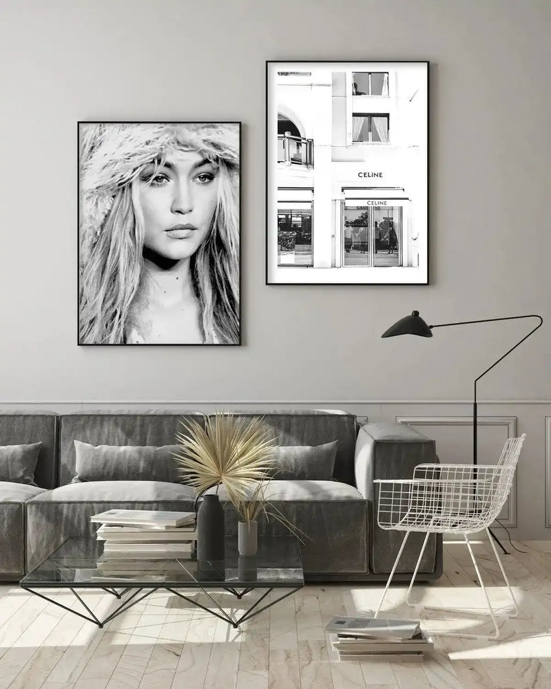 Black and white photograph art print displayed in a charcoal gray frame with white border, showing a woman's portrait with long straight hair and a fur or textured hat, positioned above a gray upholstered sofa in a modern living room next to a framed architectural line drawing of a Celine storefront. The print has a minimalist aesthetic with neutral tones throughout the space.